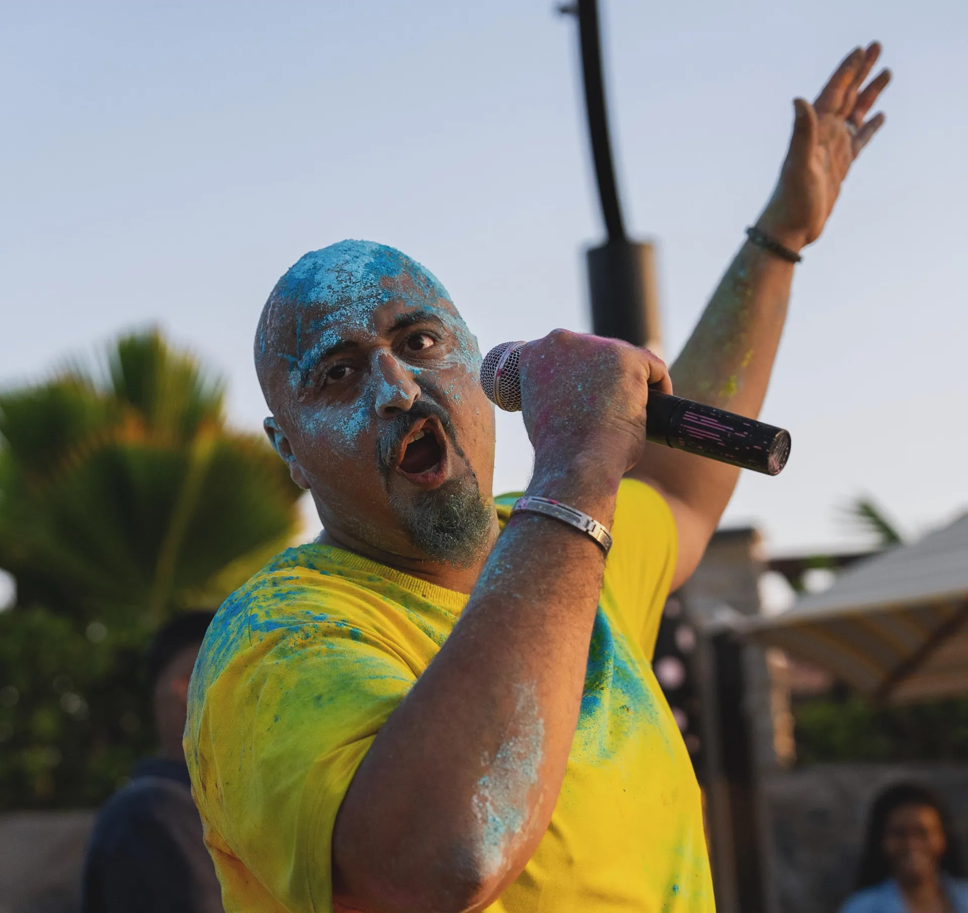 Singer mid-shout into a microphone, face dusted blue, at a beach colour festival