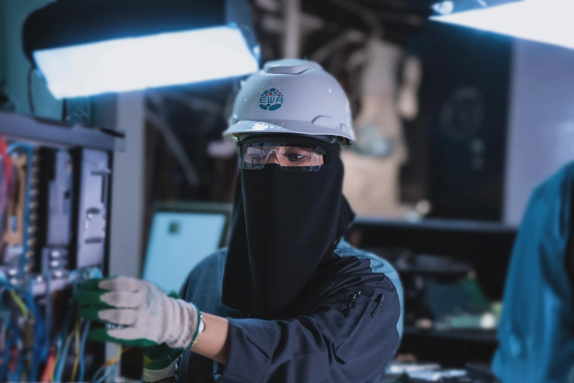 Engineer in niqab and hard hat wiring a control panel under set lights