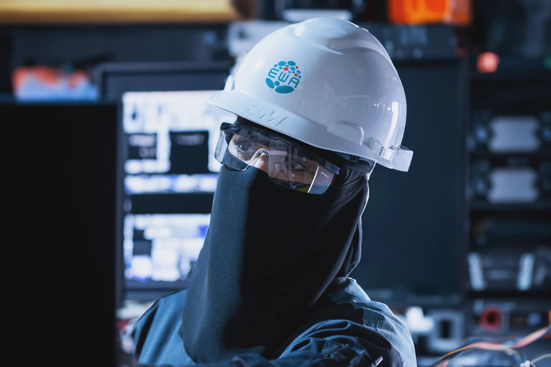 Engineer in a white hard hat and safety glasses, lit by control-room screens