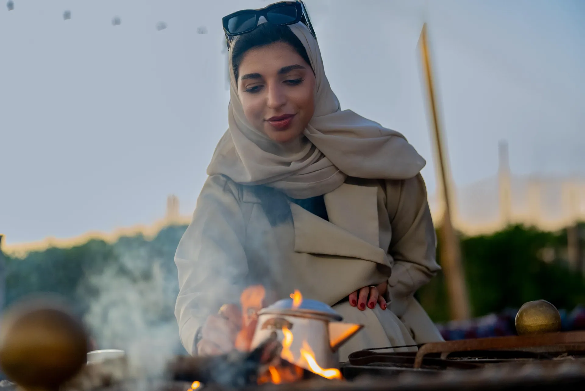 Woman in a cream headscarf brewing coffee over an open fire, smoke drifting