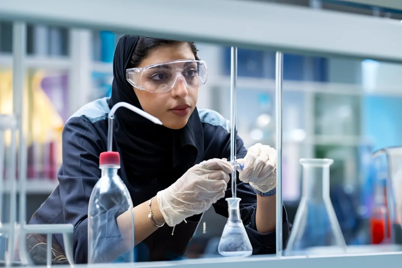 Trainee in a hijab and safety glasses measuring a sample at a lab bench