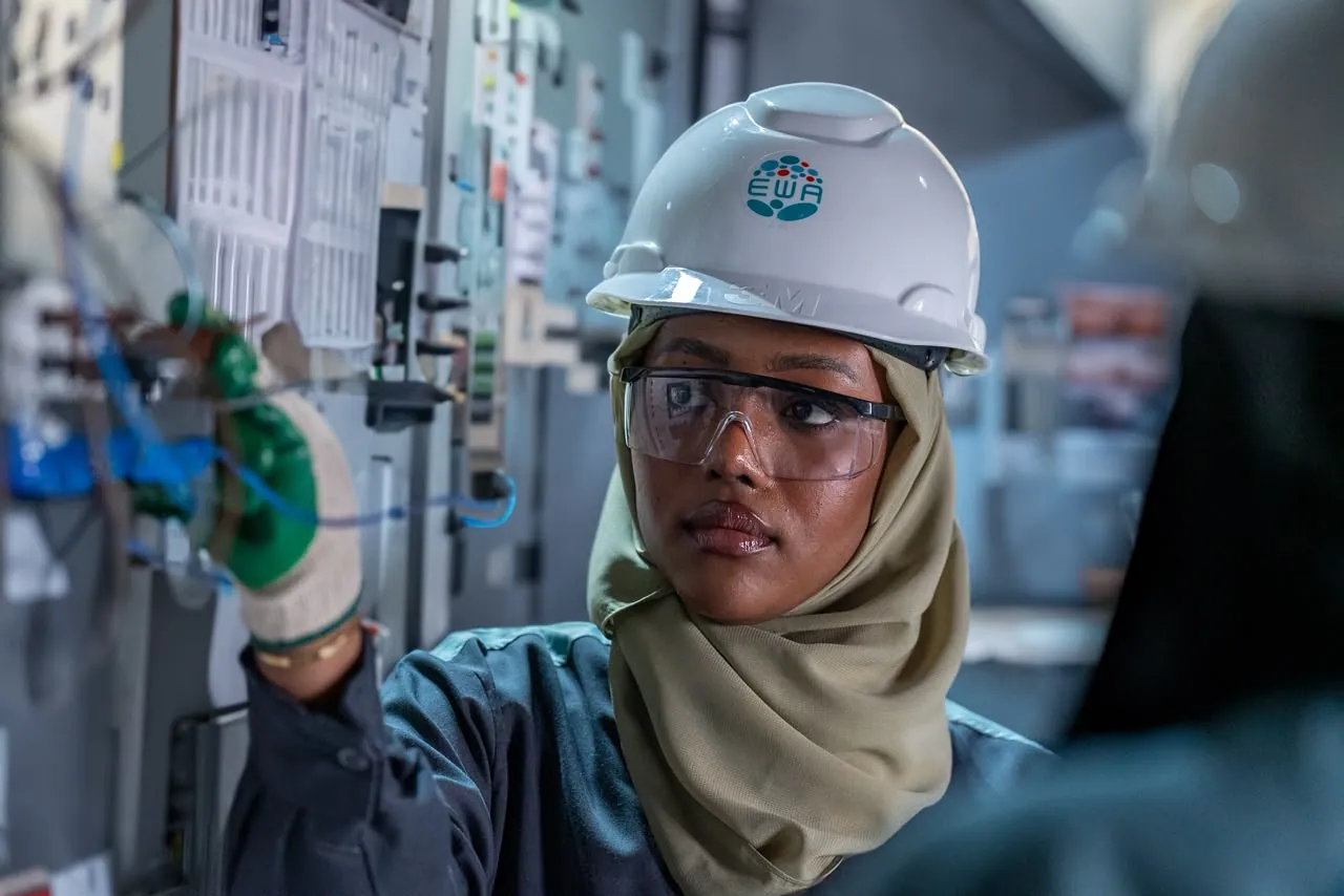 Engineer in a hijab and EWA hard hat reaching into a control panel